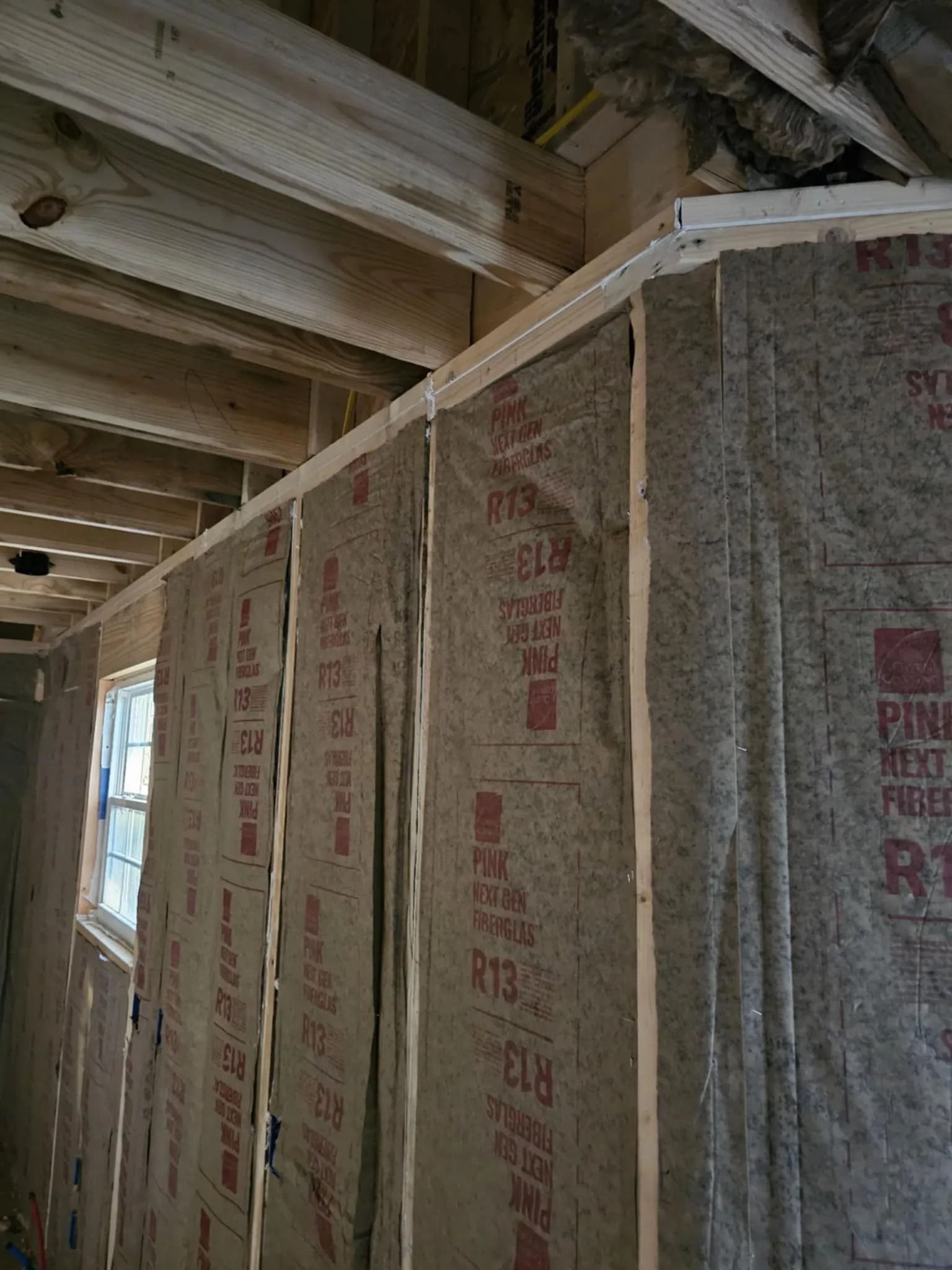 Interior of a building under construction, showing insulated wall, window, and exposed wooden ceiling beams.