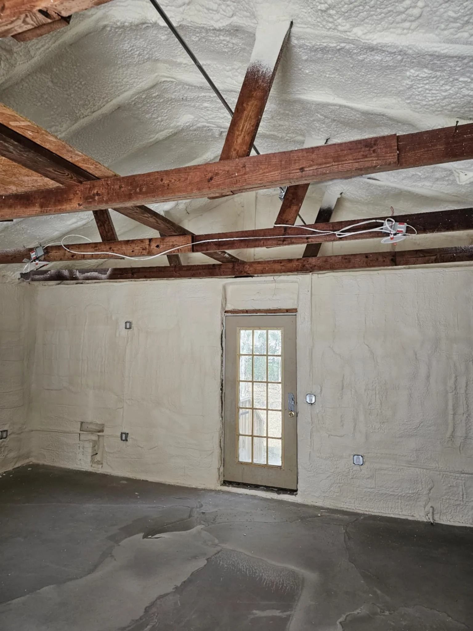 Room with spray foam insulation on ceiling and walls; door in center, concrete floor.