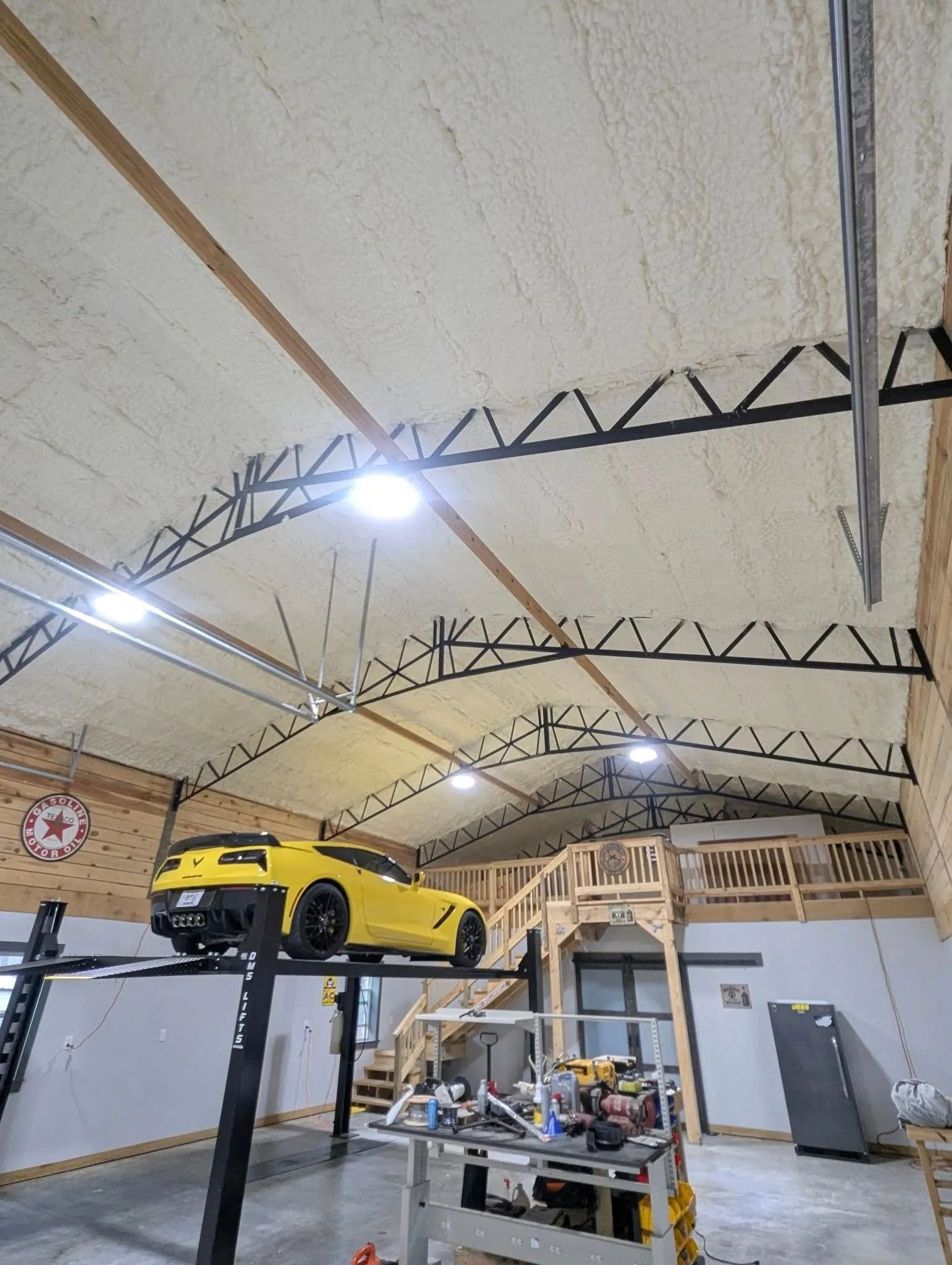 Yellow sports car on a lift in a garage with spray foam insulation, wood beams, and a mezzanine.