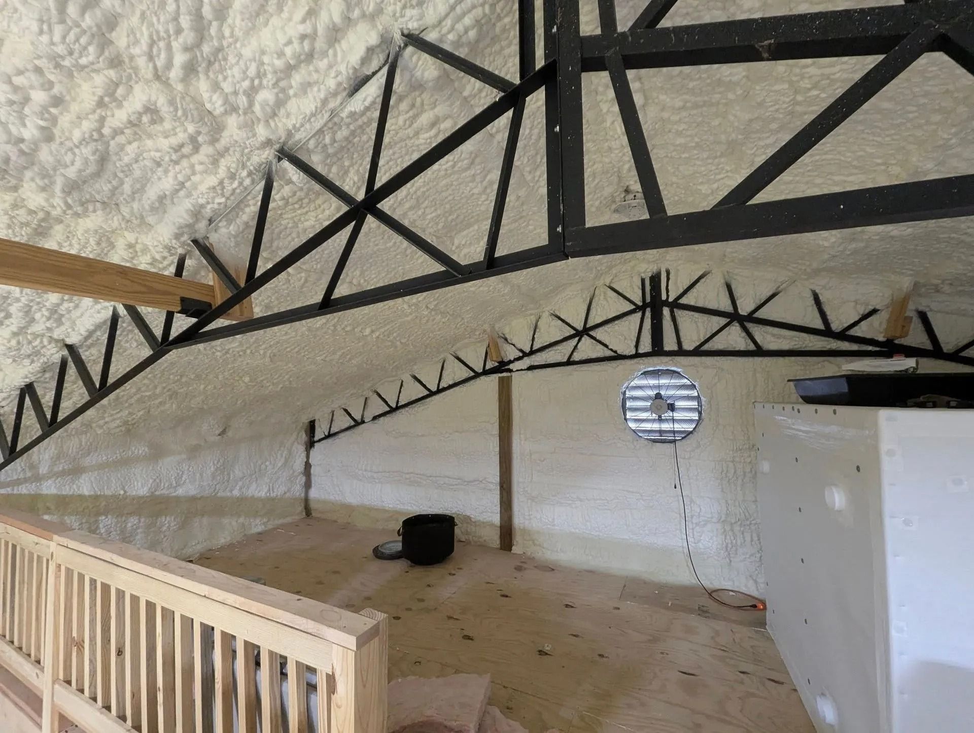 Interior view of a building's attic with exposed beams, spray foam insulation, and a partial view of a wooden railing.