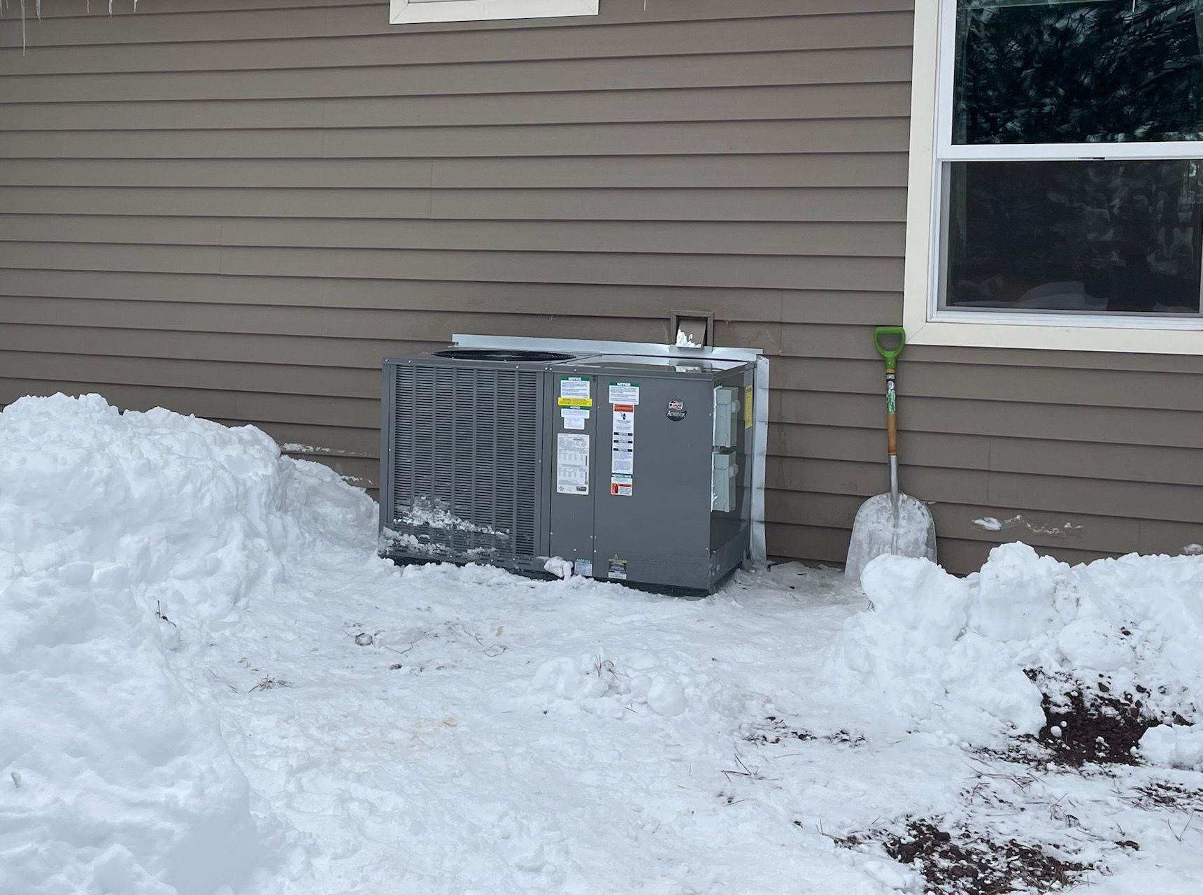 An air conditioner is sitting in the snow in front of a house.
