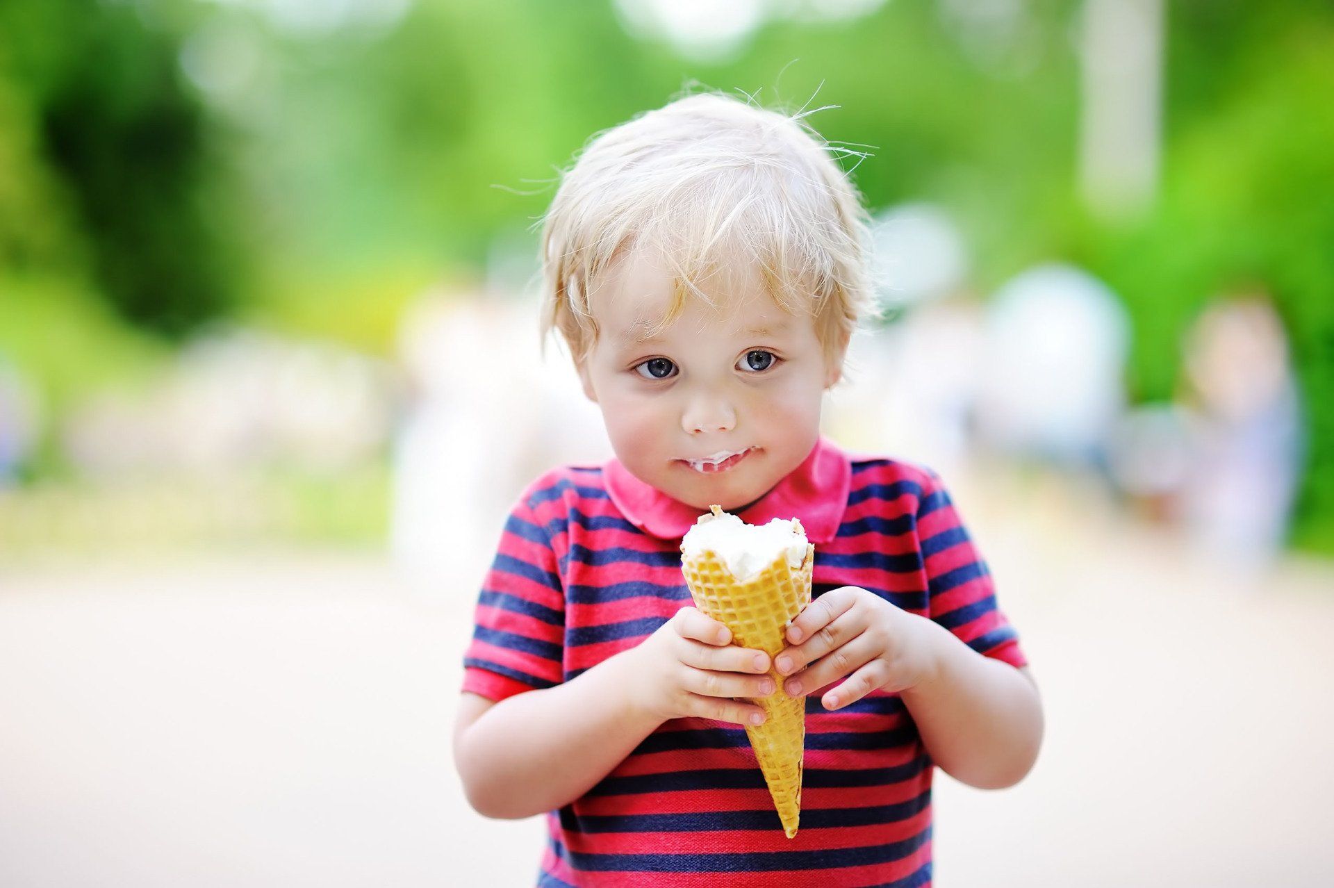 Blond child eating ice cream cone, red and blue striped shirt, outdoors, looking at camera.