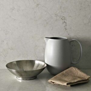 Silver bowl, white pitcher, and folded cloth on a marble-like surface against a mottled background.