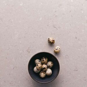 Bowl of quail eggs on a textured, light gray surface. Three eggs are outside the bowl.