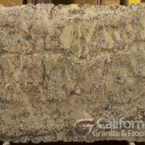 A large slab of granite, tan with grey and brown veining, displayed at a showroom. The surface has a textured, rough appearance.