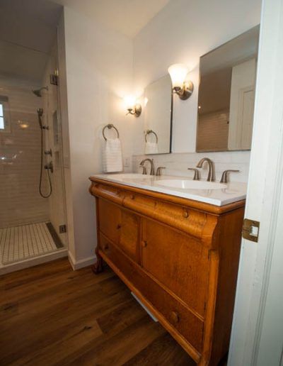 A bathroom with a wooden vanity, marble countertop, dual sinks, twin wall mirrors, and a glass-enclosed shower.