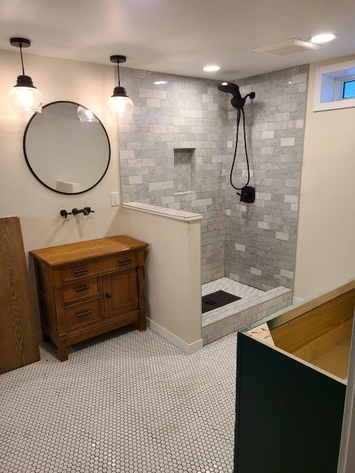 A bathroom with a white tiled floor, a wooden vanity with a round mirror, and a walk-in shower with grey brick-style tiles.