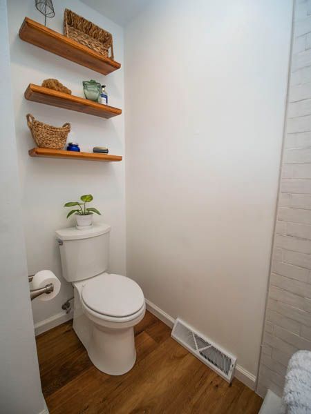 A bathroom featuring a toilet, three wooden floating shelves with decor, and wood flooring against white walls.