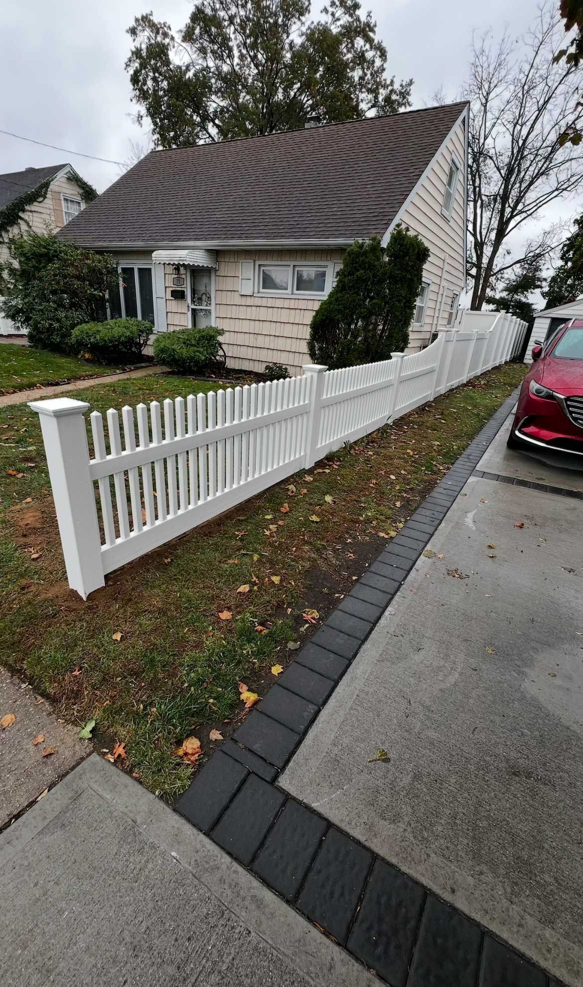 White picket fence bordering a light-colored house with a dark roof and a paved driveway.