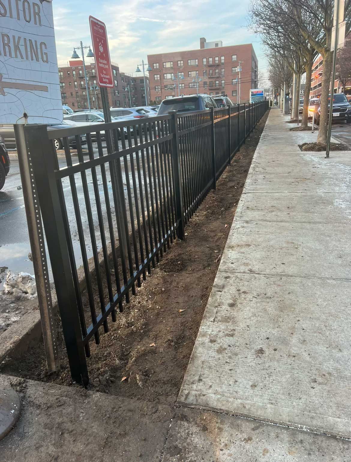 Black metal fence along a sidewalk; bare earth between the fence and pavement. Cars parked in the background.