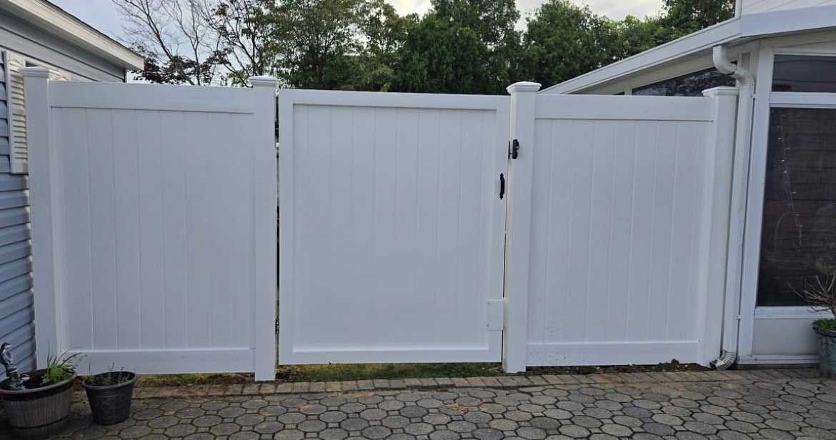 White vinyl fence with gate, set against trees and a house.