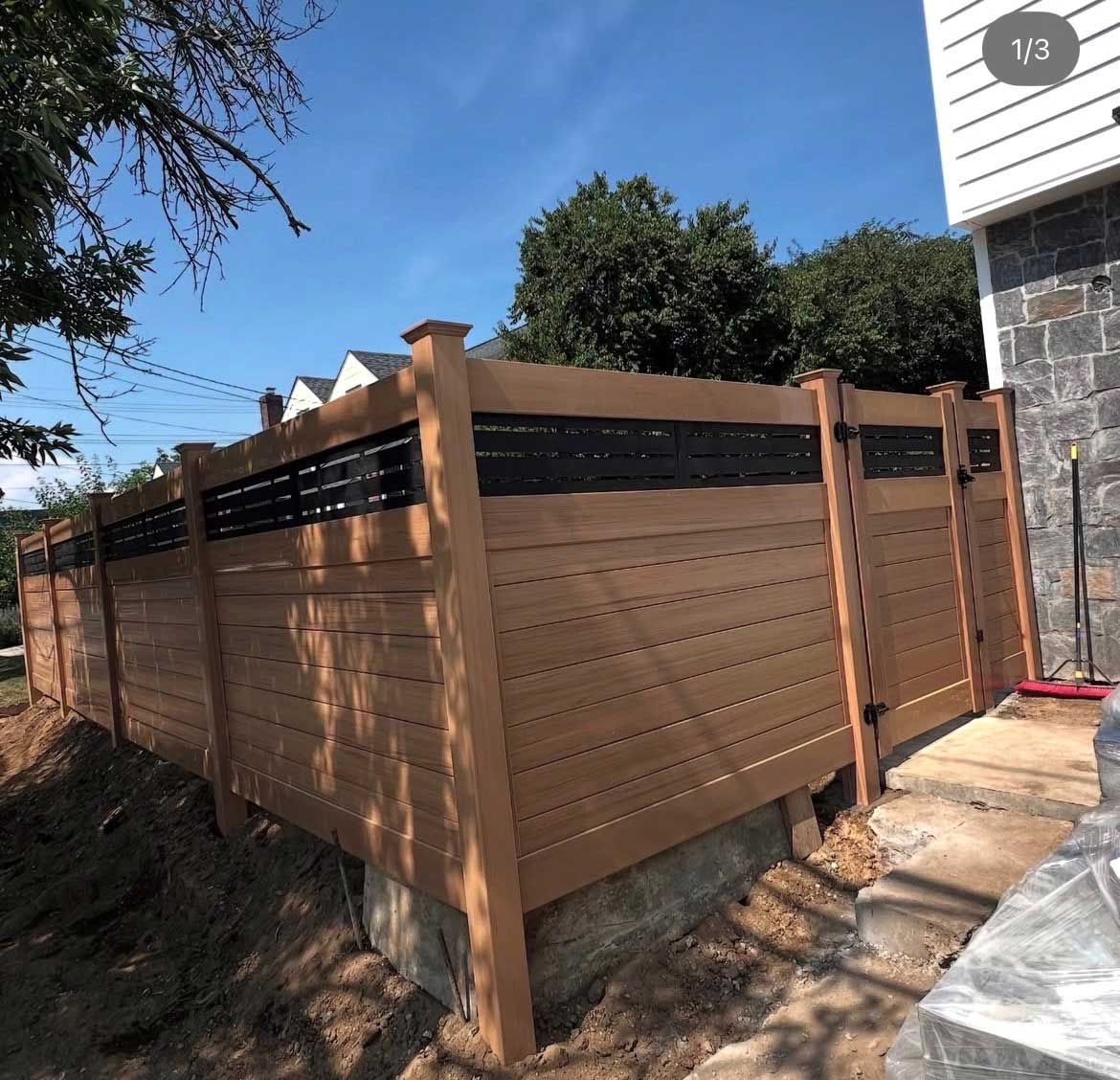Wooden privacy fence with a black top section in front of a house on a sunny day.