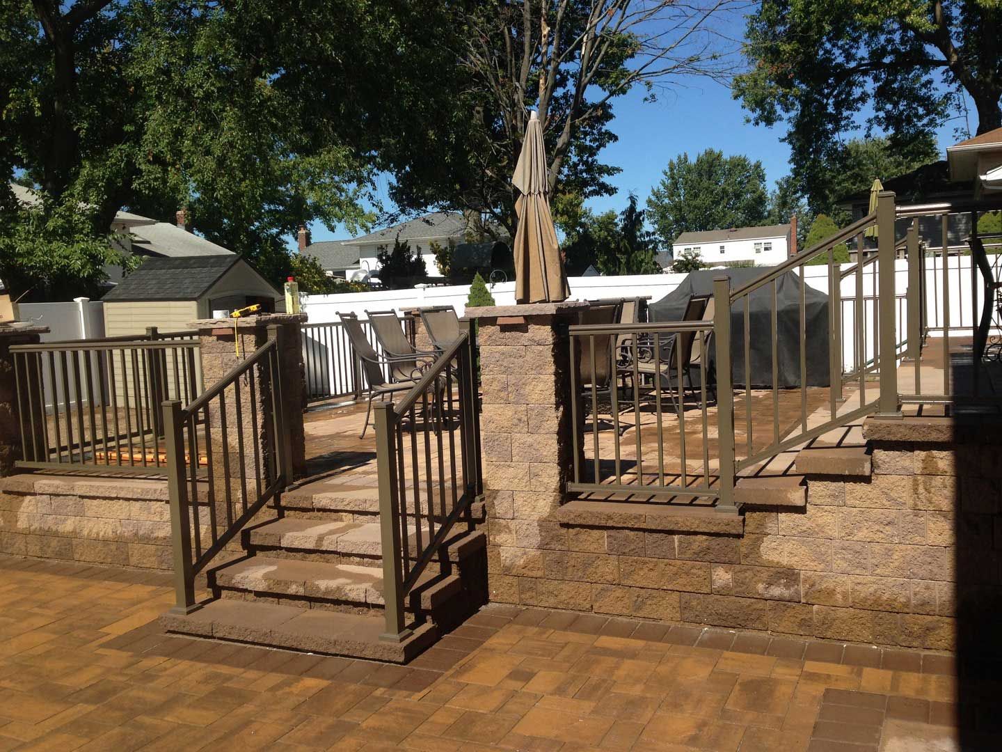 Brick patio with steps, brown metal railings, and a retaining wall. Backyard setting with trees and furniture.