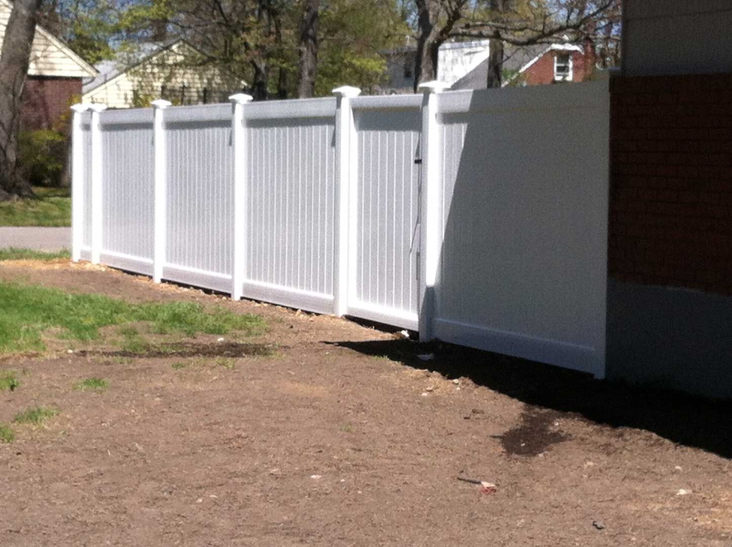 White vinyl fence with a gate, next to a brick building, and a patch of grass.
