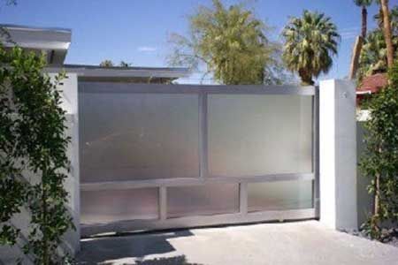 Metallic sliding gate on a driveway, flanked by white pillars and greenery.