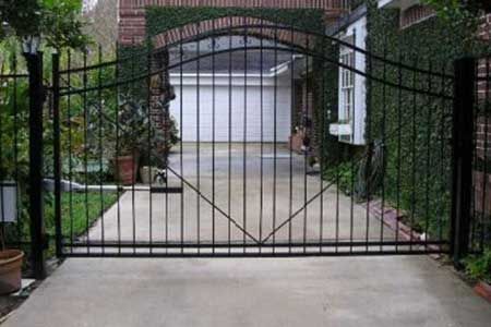Black metal gate across a concrete driveway leading to a garage. Green foliage frames the scene.