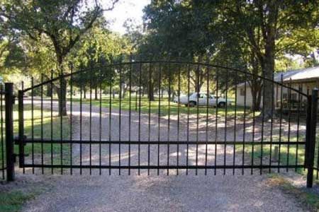 Black metal gate, arched top, blocking gravel driveway; trees and house in background.