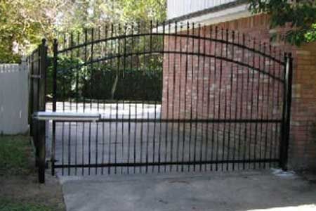 Black metal driveway gate with vertical bars and curved top, on a concrete driveway near a brick wall.