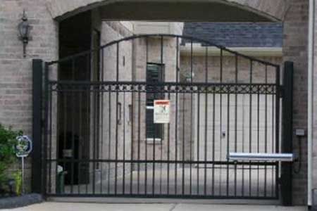 Black wrought iron driveway gate, arched top, closed, between brick pillars. Building in background.