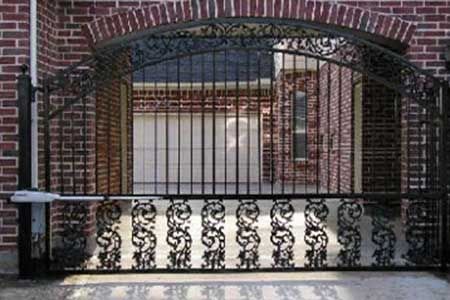 Ornate, black wrought iron gate over brick wall, automated with an electric arm, leading to a garage.