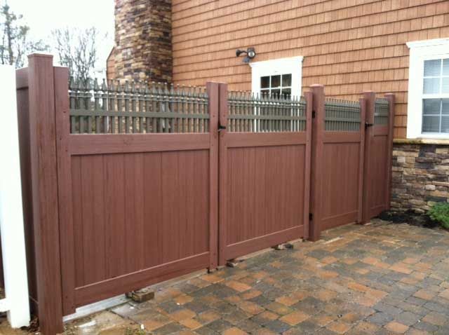 Brown privacy fence with decorative top section and brick patio in front of a house.