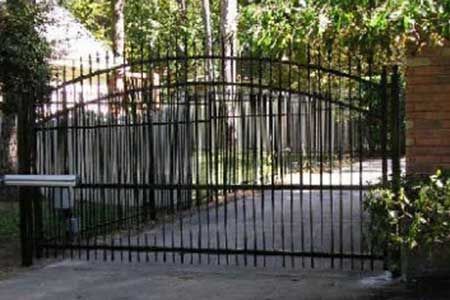 Black wrought iron gate on a concrete driveway, leading to an obscured area with trees.