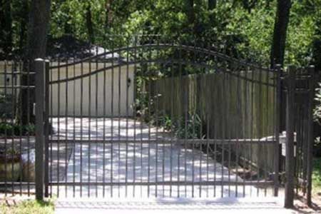Black metal driveway gate with vertical bars and arched top, set in front of a house.