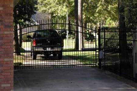 Black truck behind black metal driveway gate. Brick pillar to the left; trees and house in the background.