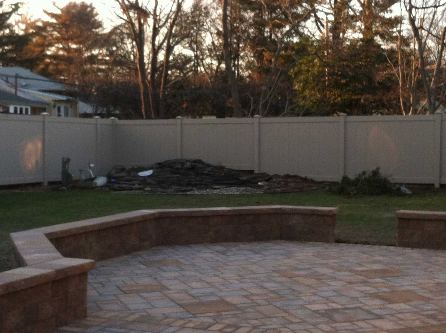 Patio with brick pavers, low wall, green lawn, beige fence, and pile of debris.