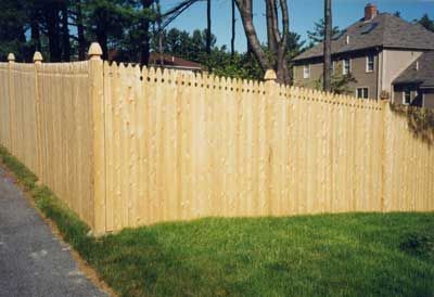 Wooden fence with decorative top surrounding a grassy yard next to a road and a house.