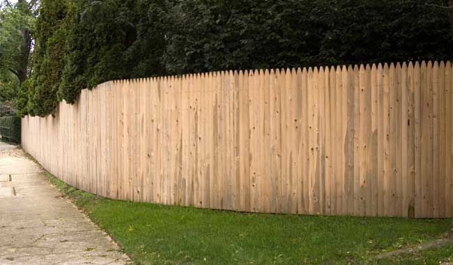 Wooden picket fence curving along a sidewalk with green grass and trees in the background.