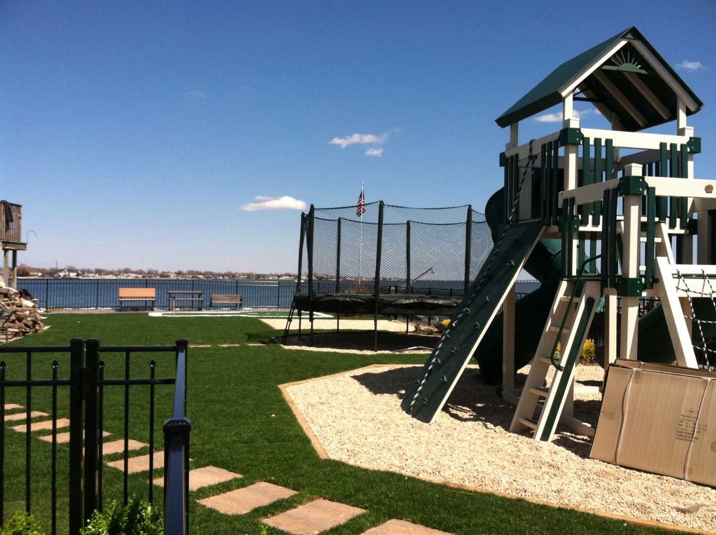 Backyard with playset, trampoline, green lawn, and water view under blue sky.