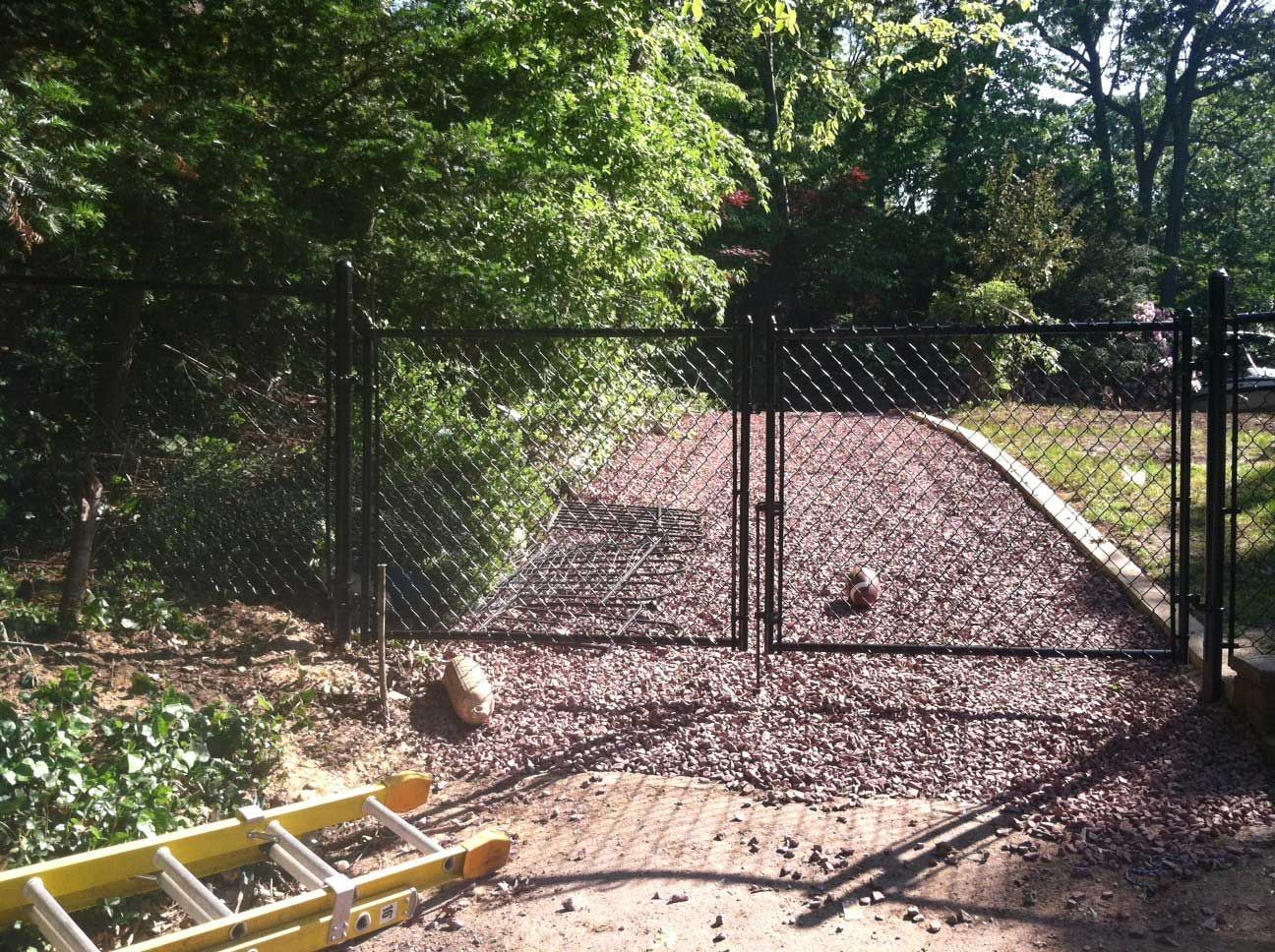 Black chain-link fence with gate, gravel pathway leading into a wooded area, yellow ladder in the foreground.