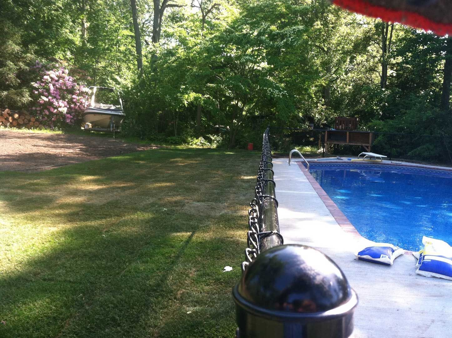 Backyard with pool, lawn, and trees. A black fence is in the foreground.