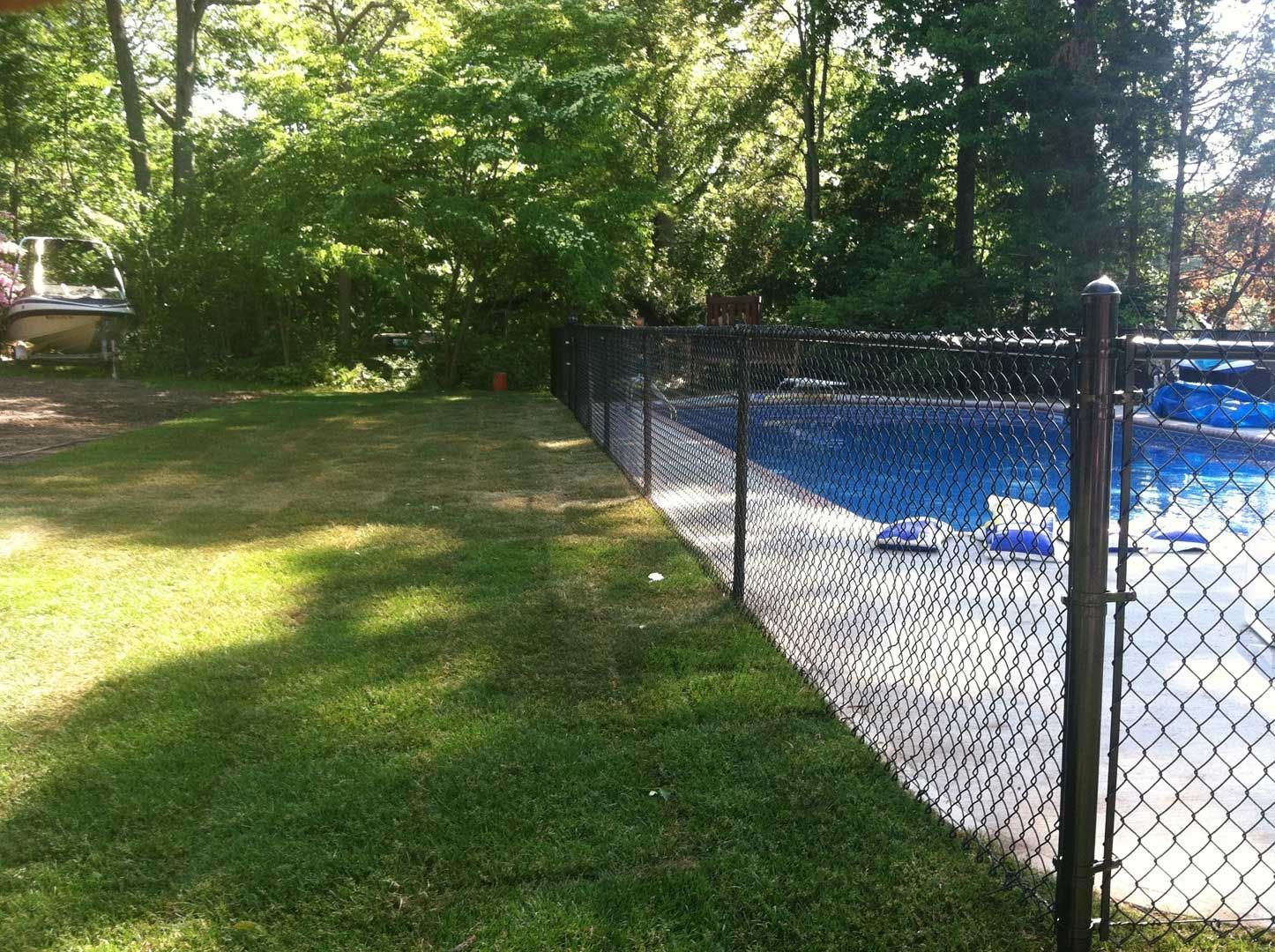 Black chain-link fence bordering a backyard swimming pool, with green grass and trees in the background.