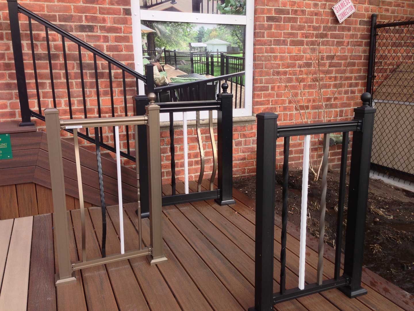 Three railing samples on a deck, against a brick wall. Each has a different post and baluster design, with variations in color.