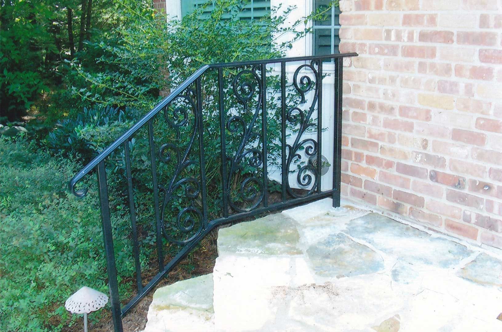Black wrought-iron railing with ornate scrollwork on stone steps next to a brick wall and bushes.