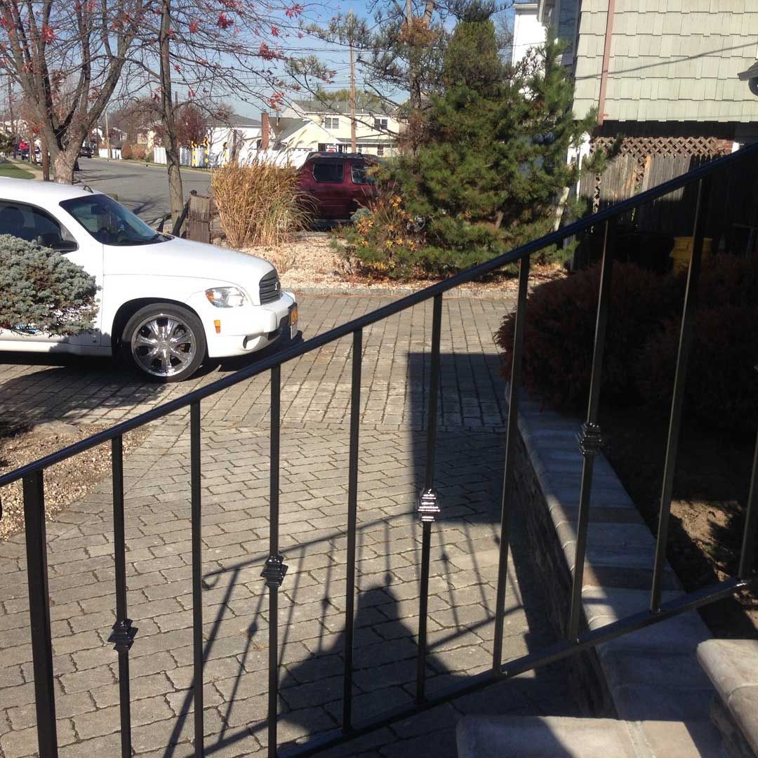 View from porch with black railing, showing driveway with white car, red SUV, and brick pavers.
