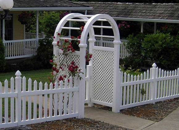 White picket fence with arched trellis gate, roses, and a house in the background.