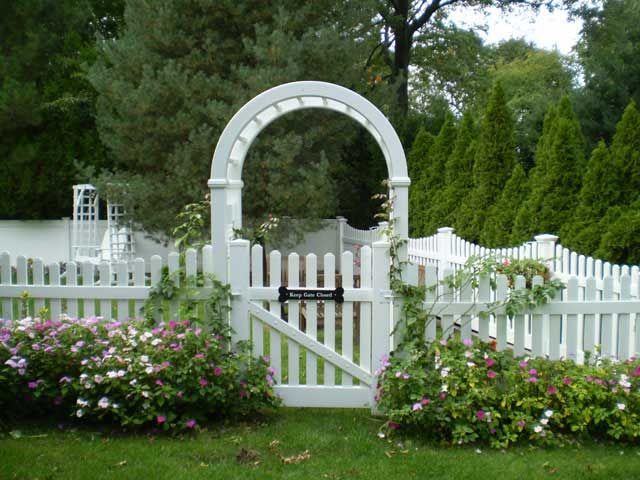 White picket fence with arched gate; roses, green lawn, trees in background.