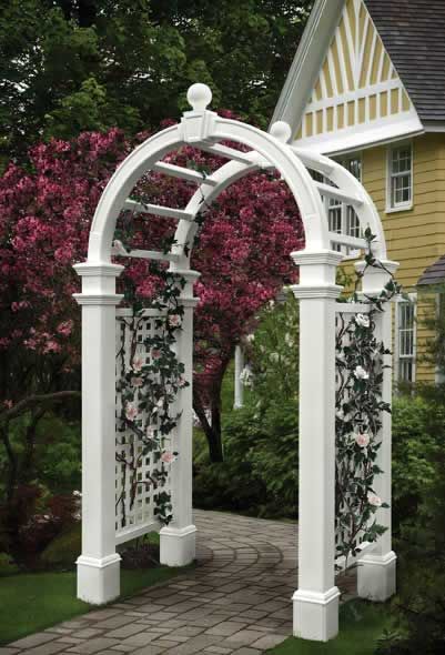 White garden arbor with climbing roses, set on a brick path, against a backdrop of trees and a yellow house.