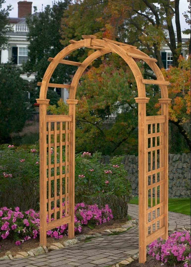 Wooden garden arbor with lattice sides, arch top, set in a garden with blooming pink flowers and brick path.
