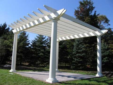 White pergola with four columns, built over a paved area, set in a grassy yard, with evergreen trees in the background.