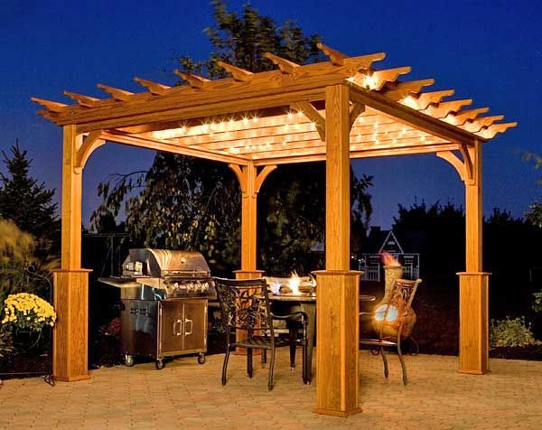 Wooden pergola with grill, table, and chairs set up for outdoor dining at night.