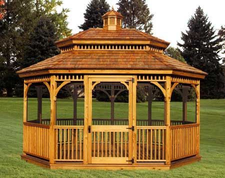 Wooden gazebo with a shingled roof, set on grass, with surrounding trees.