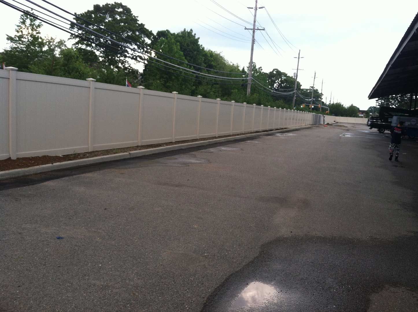 Long beige fence alongside a paved area with utility poles and trees in the background.