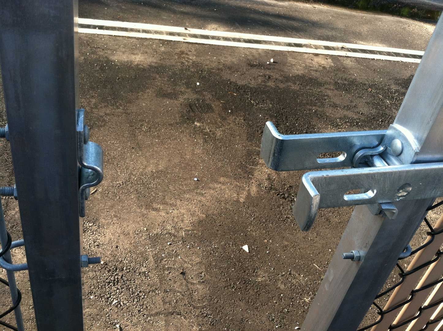 Metal gate latch on a chain-link fence, partially open. Asphalt pavement in the background.