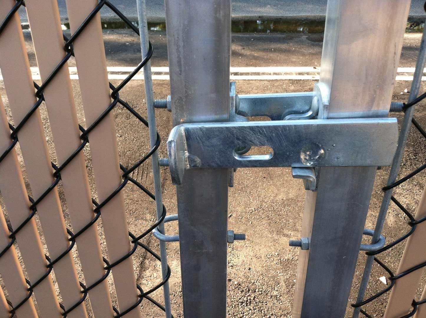 Close-up of a gate hinge attached to two metal fence posts, with tan chain link fencing visible.