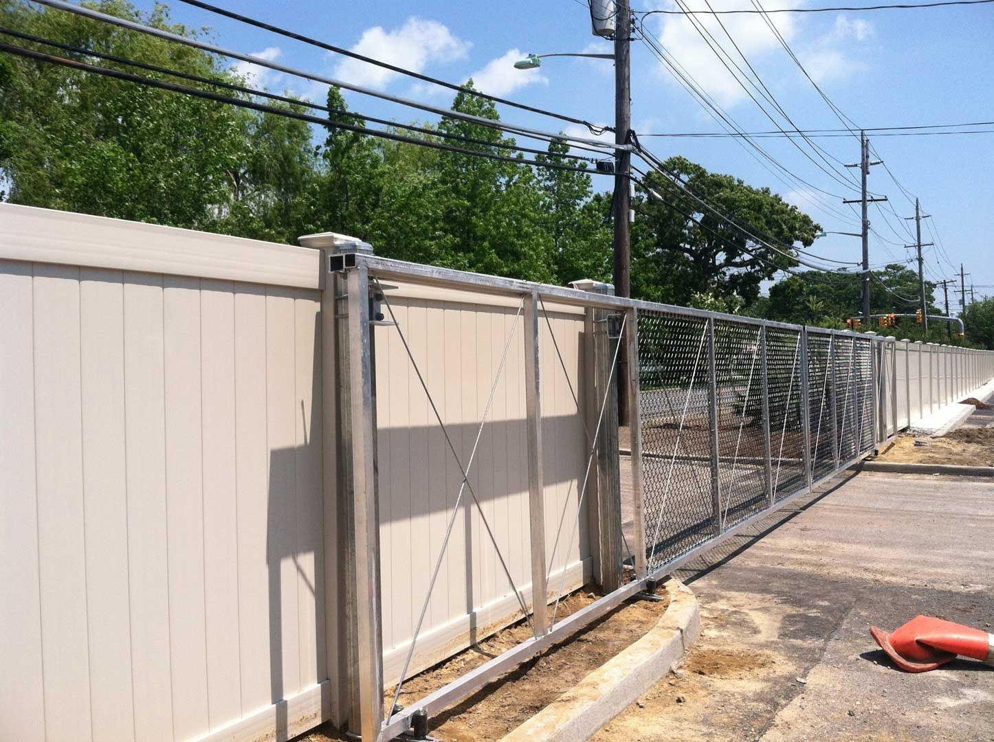 Tan vinyl fence with a metal sliding gate, outdoor setting, utility poles and green trees in the background.
