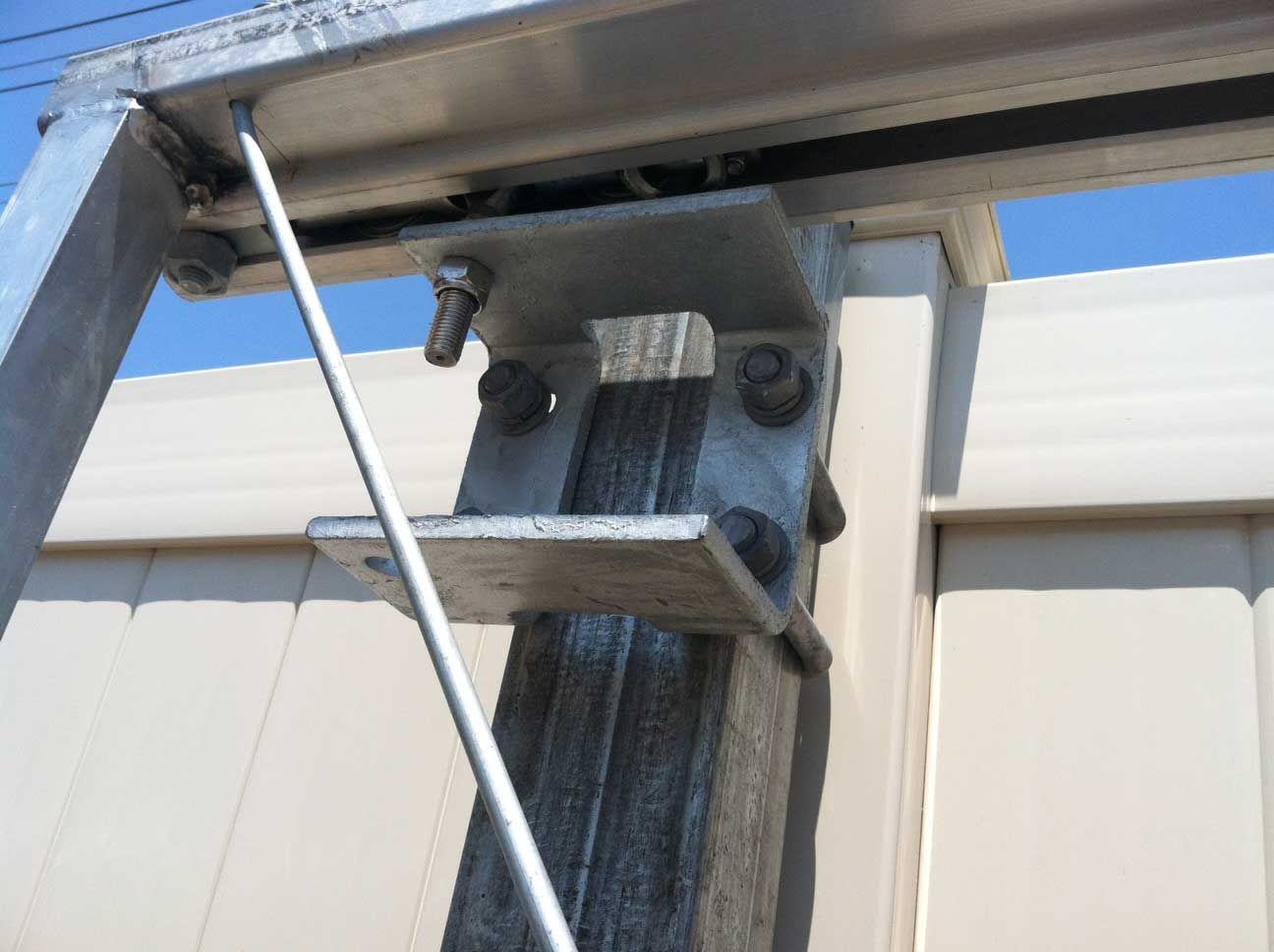 Metal gate support hardware bolted to a tan vinyl fence post and a silver overhead track.